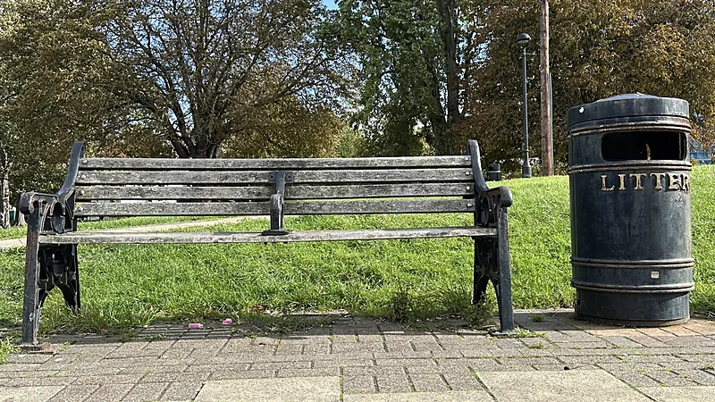 Park bench with grey, sun-bleached wooden slats on a black cast iron frame beside a litter bin
