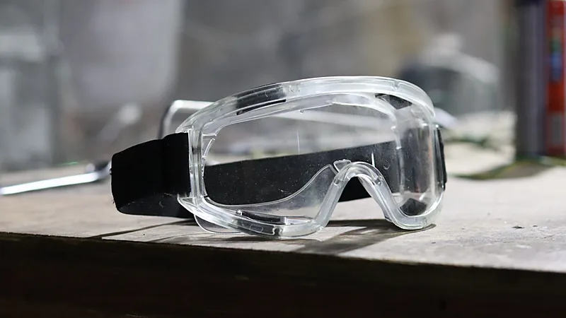 Clear safety goggles with a black elastic strap resting on a wooden workbench