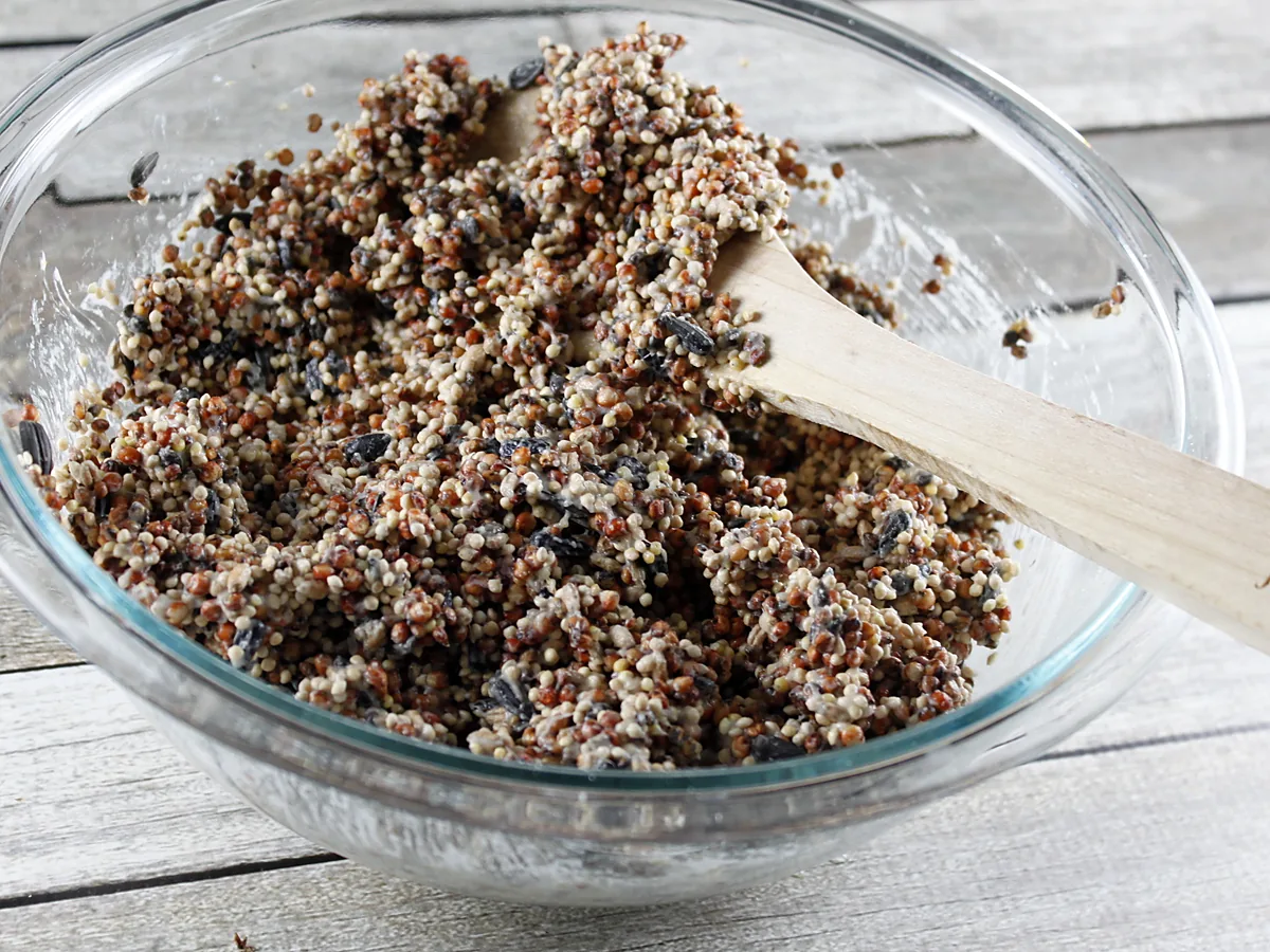A glass bowl of mixed bird seed bound with pale melted fat, stirred with a wooden spoon on a weathered wooden table