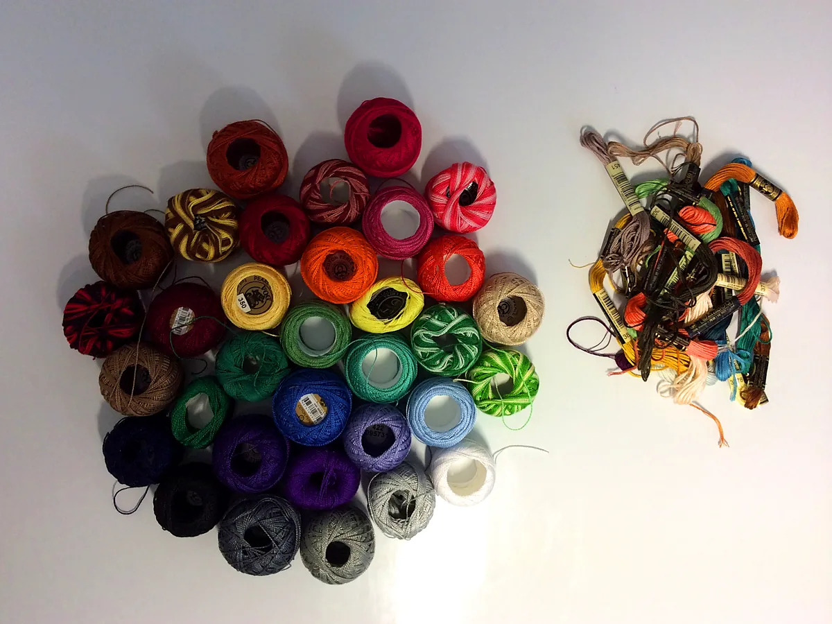 Balls of perle cotton arranged in a colour wheel on a white surface, with a tangled pile of loose thread skeins beside them
