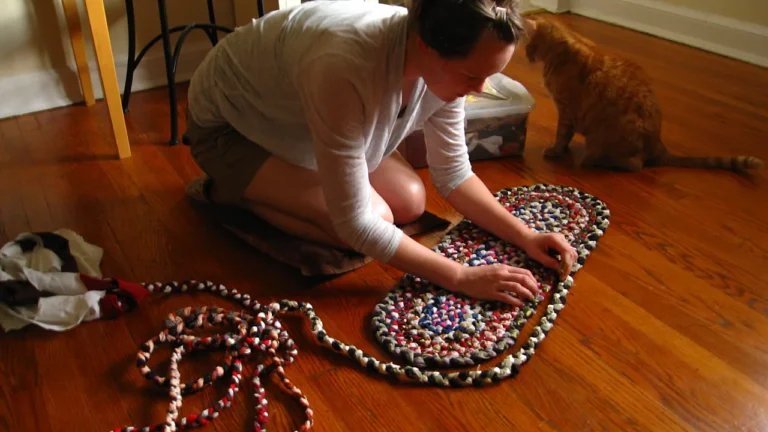 Woman kneeling on a wooden floor stitching a part finished oval braided rag rug, with a long coil of braid and a pile of old clothes beside her
