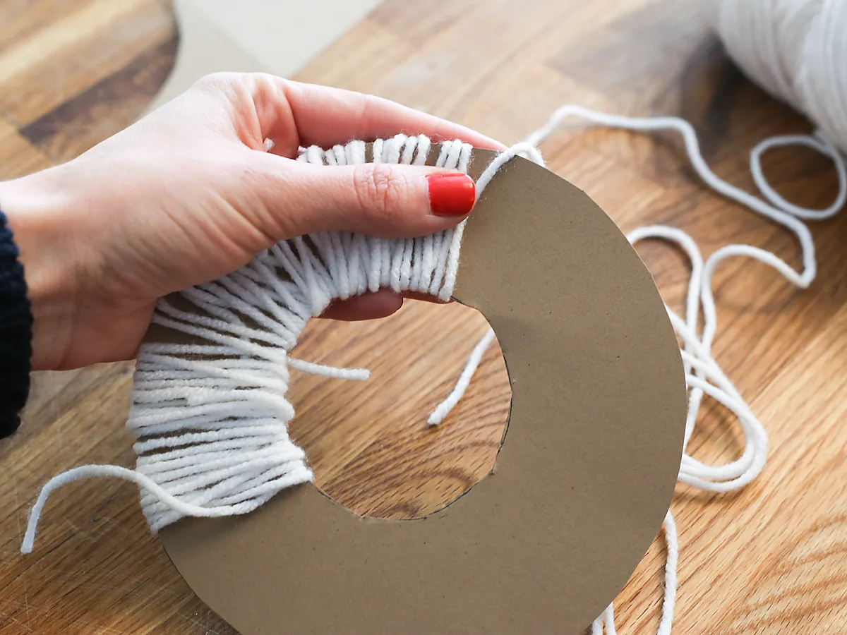 A hand winding white yarn around a doughnut-shaped cardboard ring on a wooden table, with a skein of yarn alongside