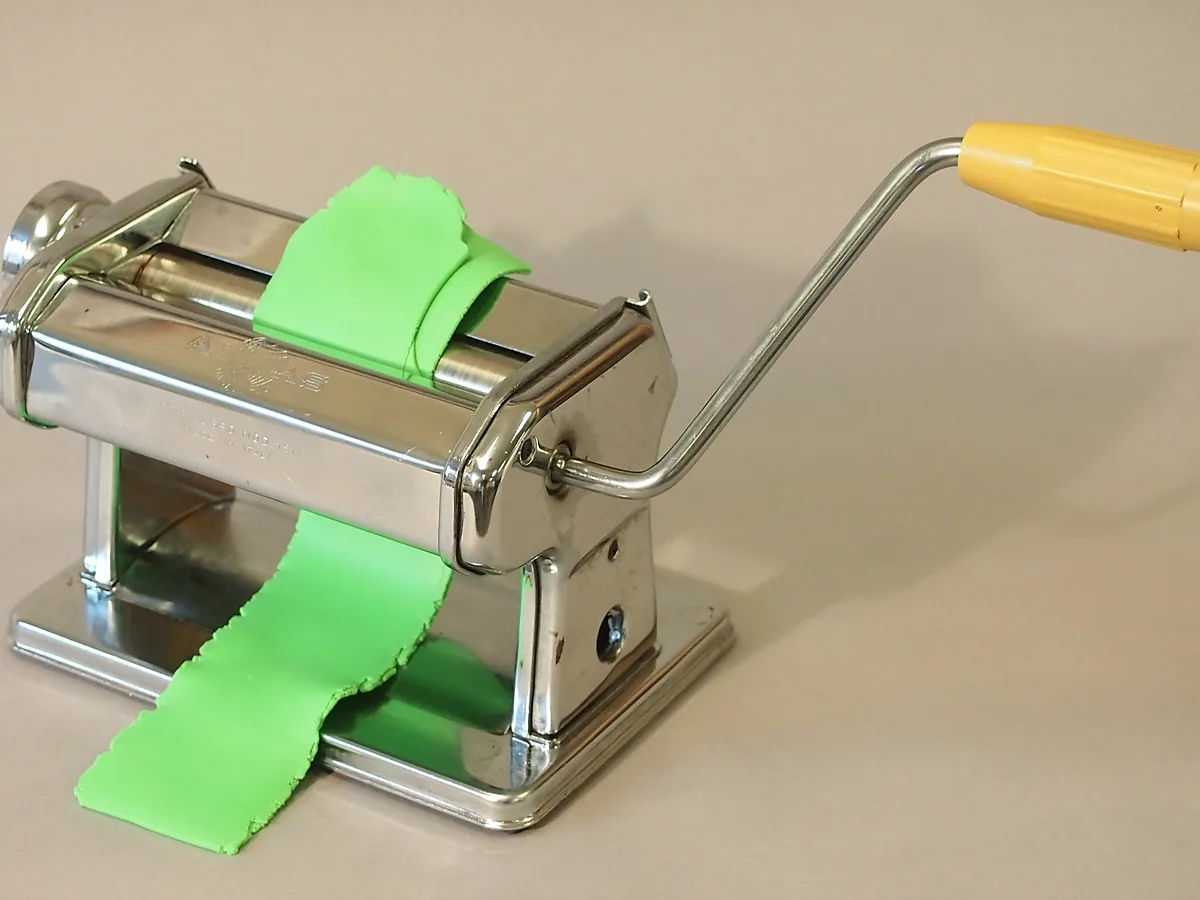 A hand-cranked pasta machine rolling a sheet of green polymer clay to an even thickness