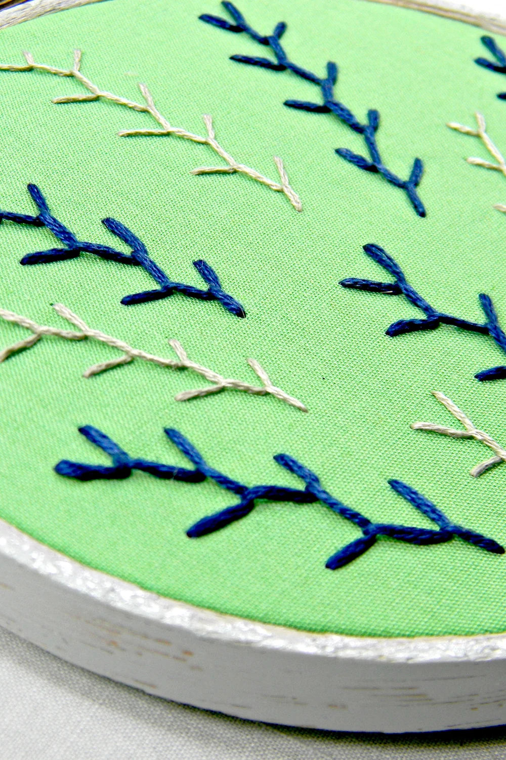 Close-up of a white wooden embroidery hoop holding green fabric stitched with rows of navy and cream feather-stitch sprigs