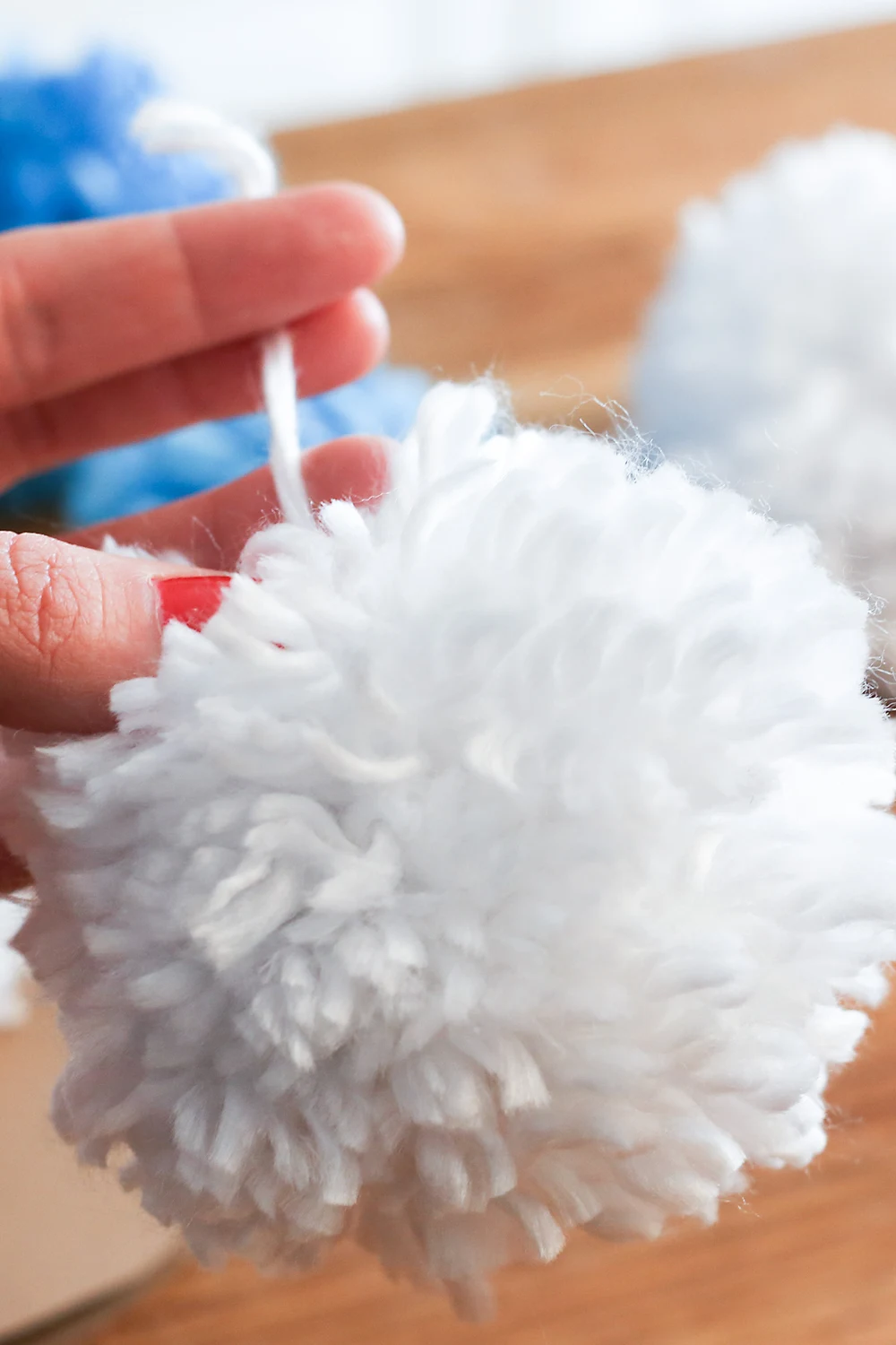 A hand holding a dense white yarn pom pom by its tie, with more white and blue pom poms on a wooden table behind