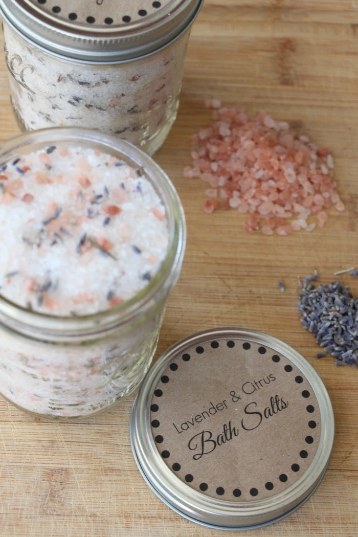Two jars of homemade bath salts with pink salt and dried lavender on a wooden board, beside a kraft paper lid labelled Lavender and Citrus Bath Salts