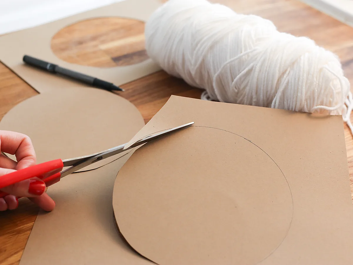 A hand cutting a large circle from brown cardboard with red-handled scissors, beside a pencil and a ball of white yarn on a wooden table