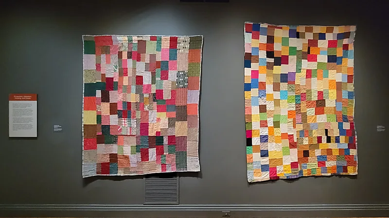 Two improvisational patchwork quilts hanging flat side by side on a dark gallery wall