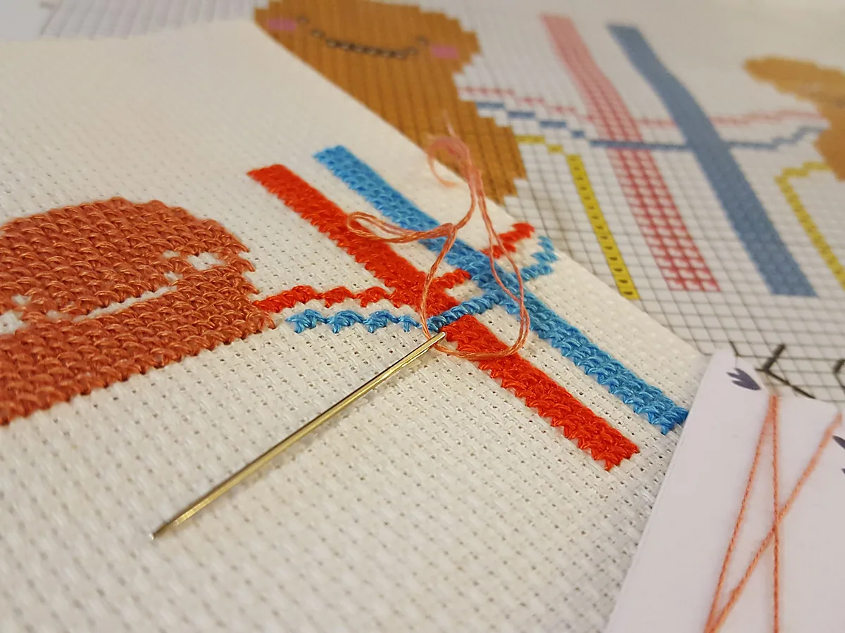 A cross stitch in progress on cream aida fabric, with a threaded needle mid-stitch and a printed chart lying alongside