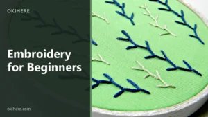 Close-up of a white wooden embroidery hoop holding green fabric stitched with navy and cream feather-stitch sprigs, with the title Embroidery for Beginners overlaid